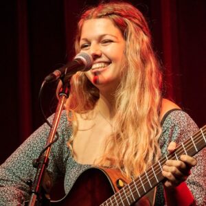 Lena Huckemann mit Gitarre im arm lächelnd am Mikrofon.