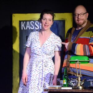 Eva Kraiss und Michael Baute stehen auf der Bühne vor einem Tisch mit Leselampe und Lektüre.