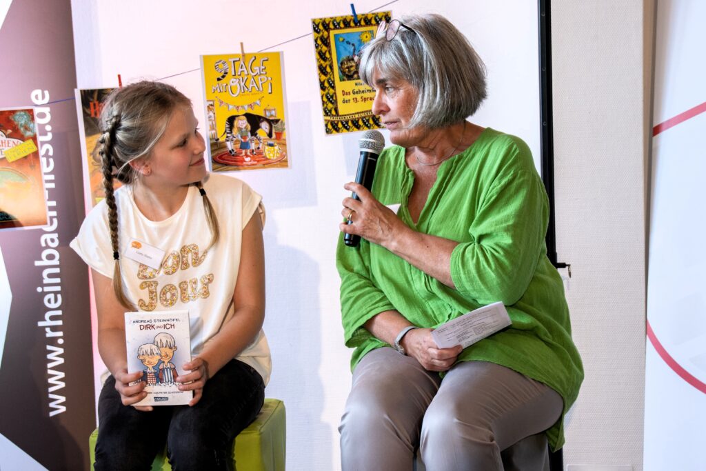 Moderatorin Elke Dietrich-Rein fragt Greta nach ihrer Lesestelle sitzend auf zwei Hockern. - Foto Ursula Petes